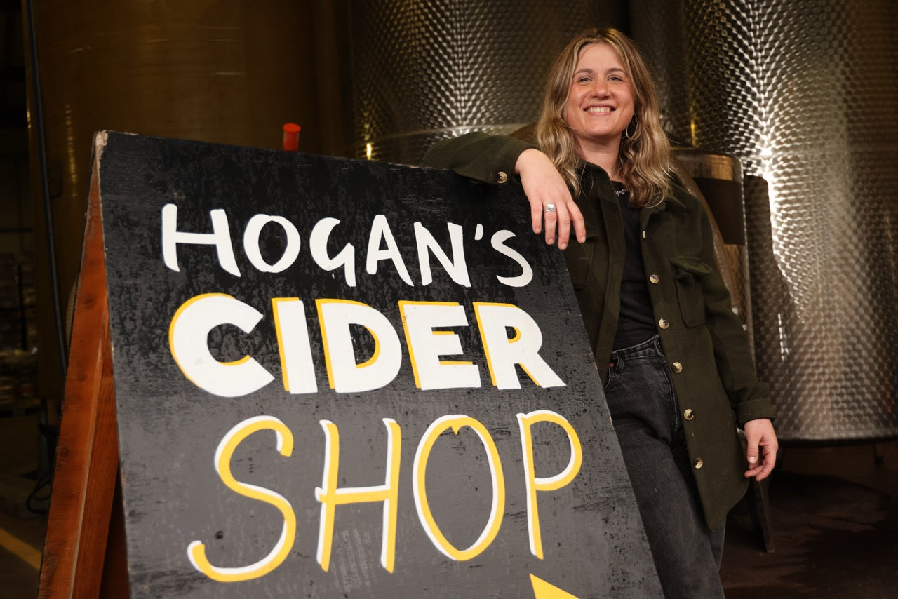 Woman holding a sign for Hogan's Cider Shop in a cider production room.
