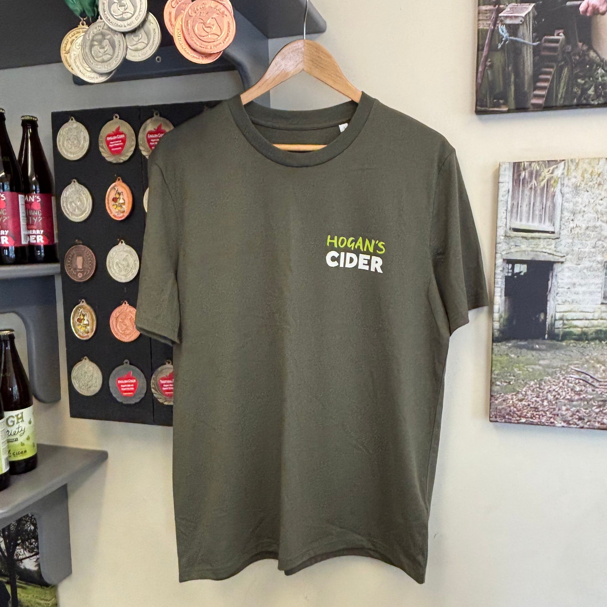 Khaki green t-shirt with 'Hogan's Cider' text on a hanger against a wall with decorative items and awards.
