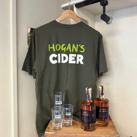 Khaki green t-shirt with 'Hogan's Cider' text on a hanger behind bottles of Hogan's Apple Brandy and Hogan's Cider branded glasses.